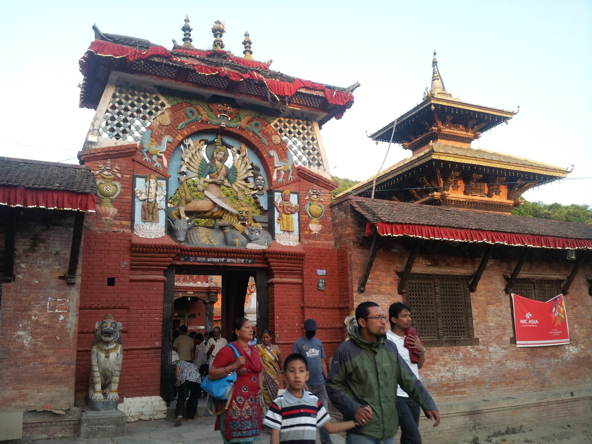 Alt text: A vibrant entrance of a traditional Nepalese temple adorned with multi-colored wood carvings of a deity surrounded by mythical creatures, flanked by stone lion statues, with people walking by and a two-tiered pagoda-style roof visible in the background.