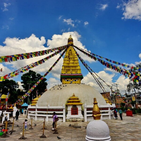 A vibrant photo of a Buddhist stupa with a golden spire, the eyes of Buddha painted on it, surrounded by colorful prayer flags radiating outward, under a blue sky with scattered clouds, with people and pigeons around the courtyard.