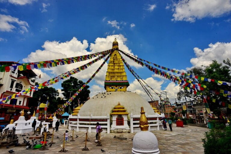 A vibrant photo of a Buddhist stupa with a golden spire, the eyes of Buddha painted on it, surrounded by colorful prayer flags radiating outward, under a blue sky with scattered clouds, with people and pigeons around the courtyard.