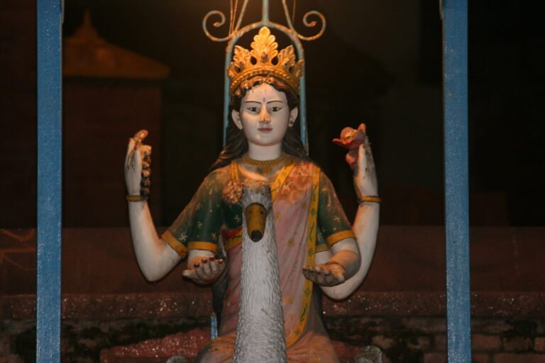 Statue of a four-armed deity with a white cow head centerpiece, holding objects in each hand, crowned and framed by a blue metallic structure, set against a shadowy background.