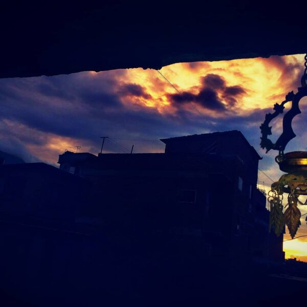 A silhouette of a hanging ornate lantern against a vibrant sunset sky with dark clouds, overlooking a silhouette of a city skyline.