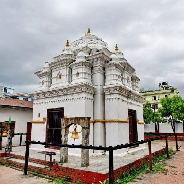 A white traditional Nepalese temple with golden accents under a cloudy sky, surrounded by a metal fence, with a front bell on a wooden frame and a building with Nepali script in the foreground.