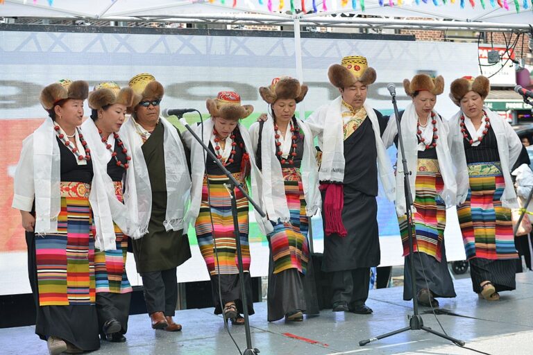 A group of people in traditional Tibetan attire performing on stage, with colorful striped aprons and white ceremonial scarves.