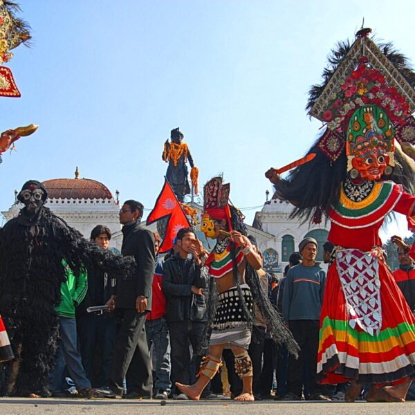 Participants in traditional costumes and masks perform a cultural dance during a festival in Nepal, with onlookers and Nepali flags in the background.