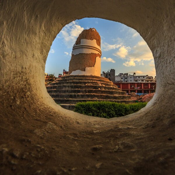 A view of a partially destroyed ancient stupa, seen through a circular hole in a wall, with buildings and a blue sky in the background.