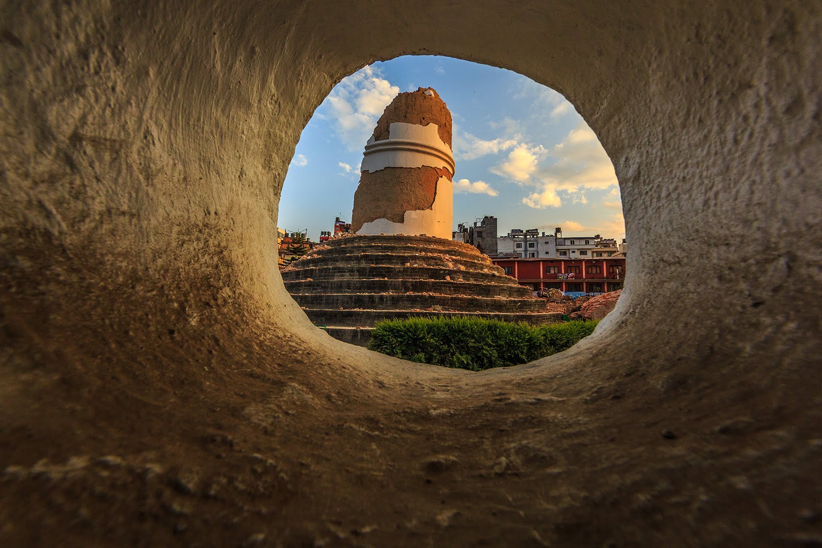 A view of a partially destroyed ancient stupa, seen through a circular hole in a wall, with buildings and a blue sky in the background.