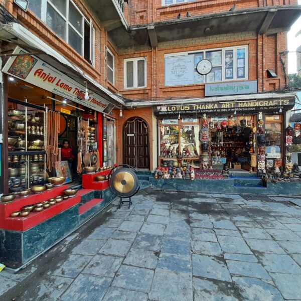 A vibrant street view showcasing two ethnic handicraft shops with displayed wares such as singing bowls, statues, and prayer beads, alongside a paved walkway.