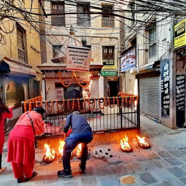 People participating in a street ritual with small fires outside a temple in a crowded alley, tangled electrical wires overhead, and closed storefronts with signage in an urban setting.