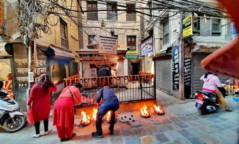 People participating in a street ritual with small fires outside a temple in a crowded alley, tangled electrical wires overhead, and closed storefronts with signage in an urban setting.