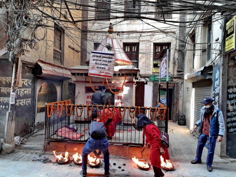 A bustling street scene with people walking by small fires in front of a Hindu temple adorned with marigold garlands, set against a backdrop of densely clustered wires and aged buildings.