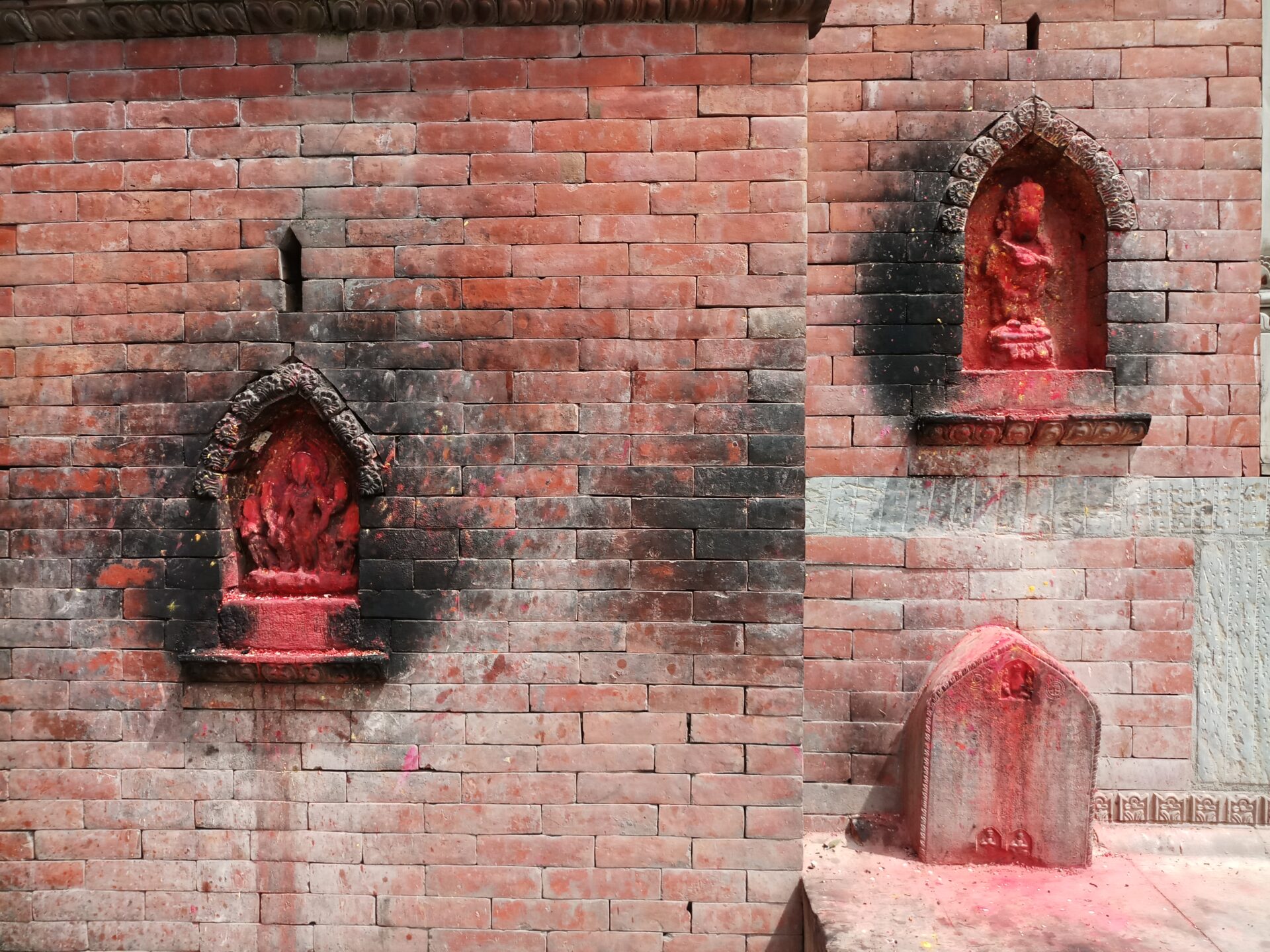 Three religious shrine statues with red pigment set into alcoves of a brick wall, exhibiting signs of worship.