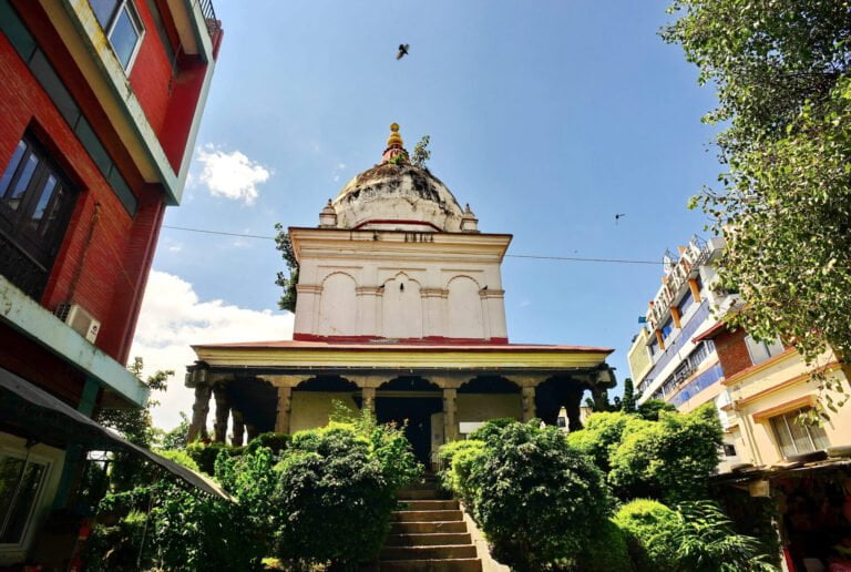 Alt text: A white domed temple with a golden spire flanked by greenery and buildings under a bright blue sky with some clouds. A flight of stairs leads up to the temple entrance.