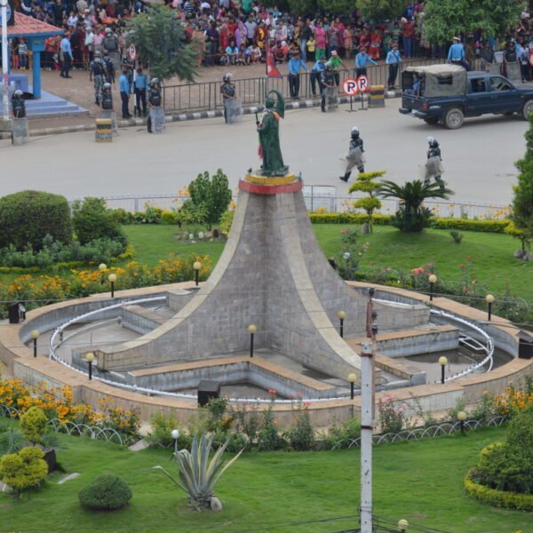 Aerial view of a public square with a central monument, landscaped gardens, and crowds of people near a perimeter cordoned off by security forces.
