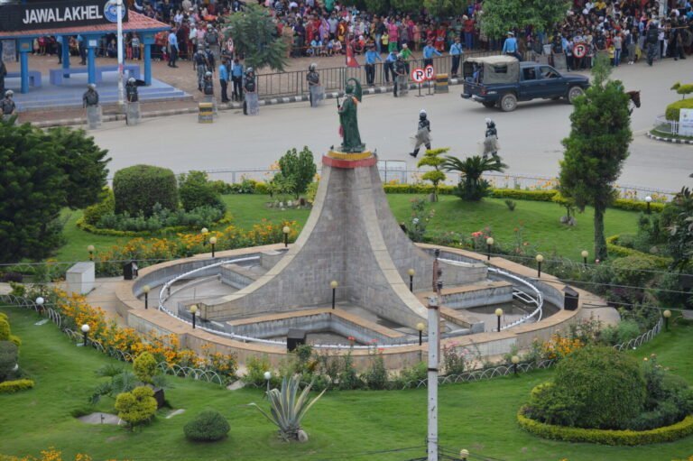 Aerial view of a public square with a central monument, landscaped gardens, and crowds of people near a perimeter cordoned off by security forces.