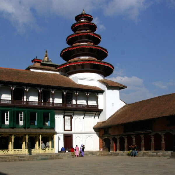 A multi-tiered traditional pagoda-style temple next to a historic building with a green balcony, set against a blue sky with scattered clouds, at Kathmandu Durbar Square, with people sitting and walking around the courtyard.