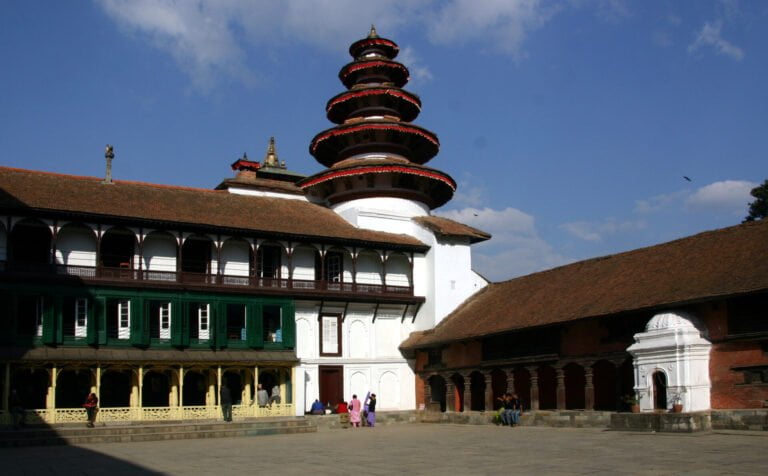 A multi-tiered traditional pagoda-style temple next to a historic building with a green balcony, set against a blue sky with scattered clouds, at Kathmandu Durbar Square, with people sitting and walking around the courtyard.