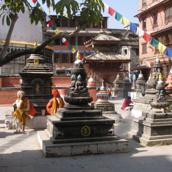 Alt text: A vibrant scene at a Nepalese temple complex with several small stone stupas in a row. Colorful prayer flags stretch above while people in traditional attire sit and converse, with a large white stupa partially visible on the left.