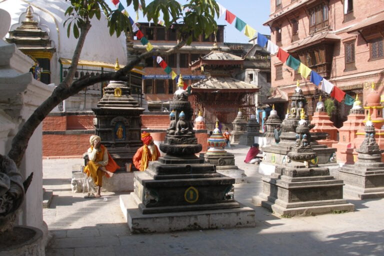 Alt text: A vibrant scene at a Nepalese temple complex with several small stone stupas in a row. Colorful prayer flags stretch above while people in traditional attire sit and converse, with a large white stupa partially visible on the left.