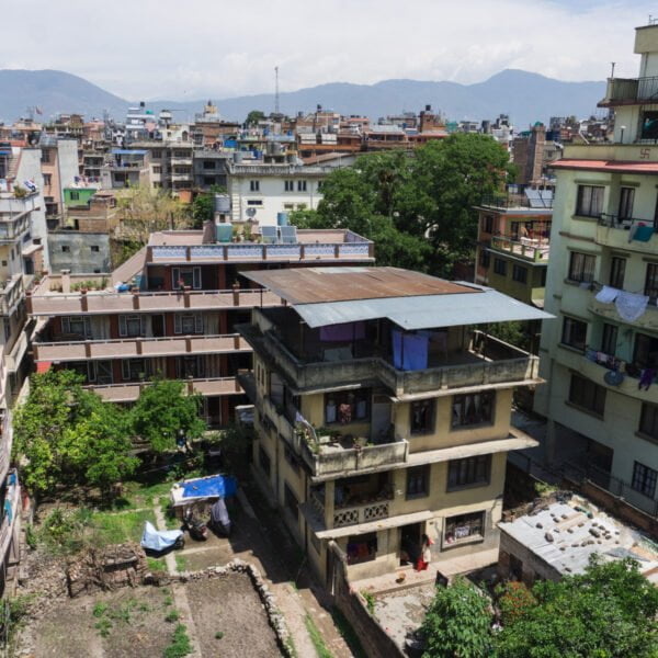 Aerial view of a densely populated urban area with multi-story residential buildings, some with clothes hanging to dry on balconies, and a patch of green space with trees surrounded by the structures, with mountains visible in the distance under a partly cloudy sky.