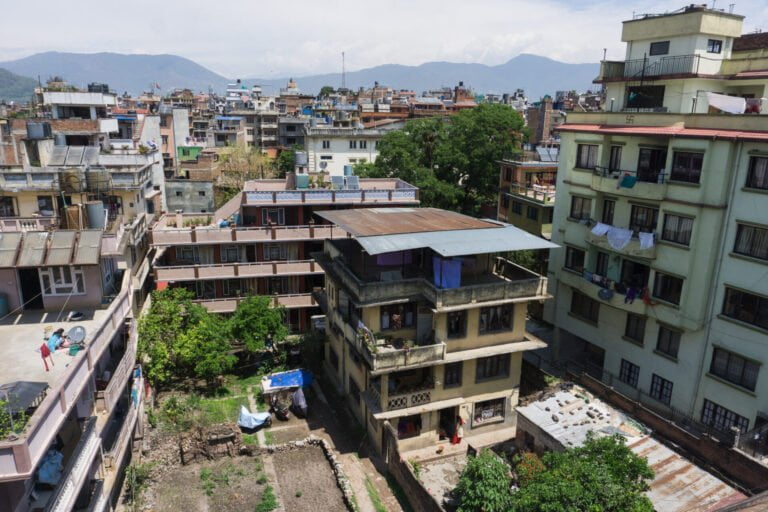 Aerial view of a densely populated urban area with multi-story residential buildings, some with clothes hanging to dry on balconies, and a patch of green space with trees surrounded by the structures, with mountains visible in the distance under a partly cloudy sky.
