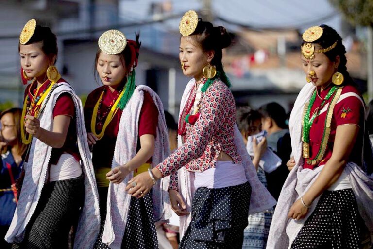 A group of four women dressed in traditional attire with gold headpieces, large earrings, and beaded necklaces performing a dance or participating in a cultural procession.