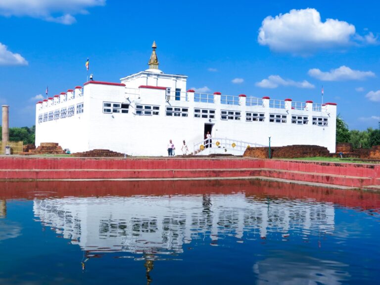 White traditional building with a gold spire reflecting in water, under a blue sky with white clouds, and visitors nearby.