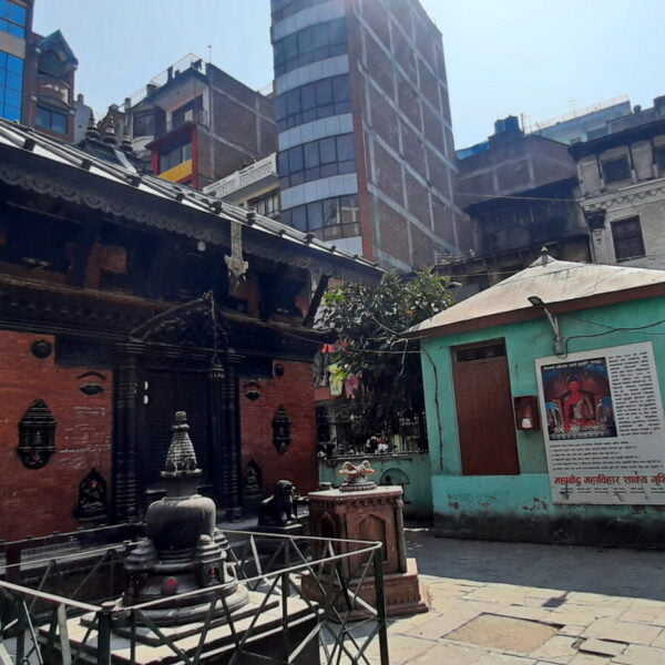 A traditional Nepalese temple with intricate wood carvings surrounded by modern buildings, a small shrine in the foreground, and people walking around in a courtyard.