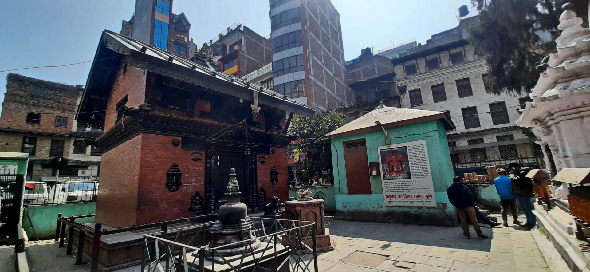 A traditional Nepalese temple with intricate wood carvings surrounded by modern buildings, a small shrine in the foreground, and people walking around in a courtyard.