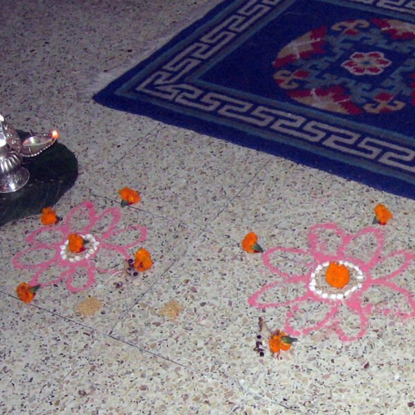 A traditional Indian floor decoration, or rangoli, made of colorful powders and adorned with flowers, with a lit oil lamp atop a stone next to it, on a terrazzo floor beside a blue rug with Greek key motifs.