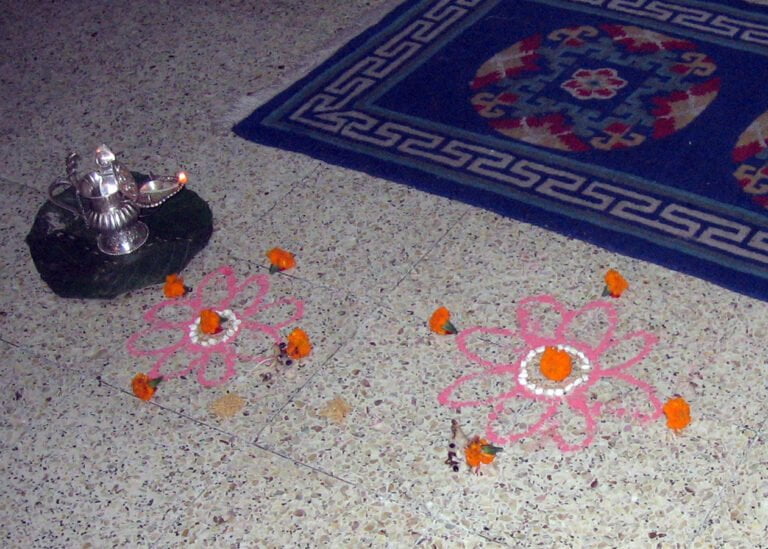 A traditional Indian floor decoration, or rangoli, made of colorful powders and adorned with flowers, with a lit oil lamp atop a stone next to it, on a terrazzo floor beside a blue rug with Greek key motifs.