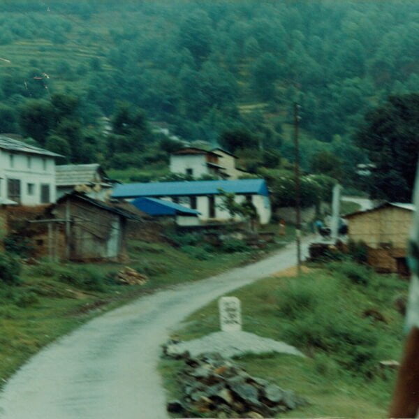 A blurry photograph showing a rural road leading through a small village with houses and forested hills in the background, and part of a person's body visible on the right edge.