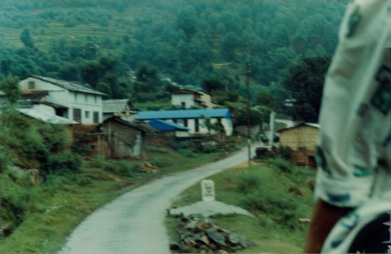 A blurry photograph showing a rural road leading through a small village with houses and forested hills in the background, and part of a person's body visible on the right edge.