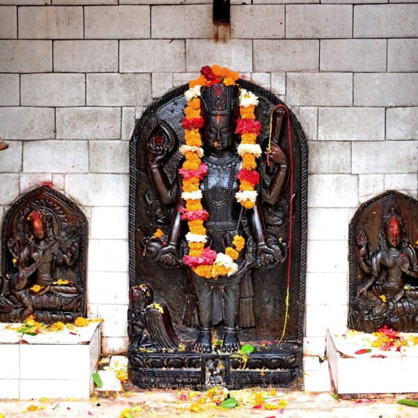 Alt text: A Hindu shrine with a central black stone statue of a deity adorned with orange and yellow marigold garlands, flanked by two smaller statues, surrounded by scattered flower petals, against a grey brick wall with a hanging brass bell.