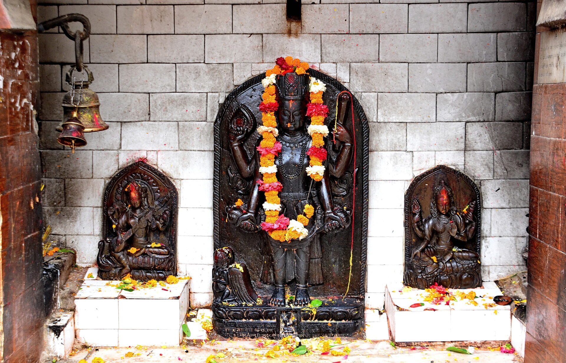 Alt text: A Hindu shrine with a central black stone statue of a deity adorned with orange and yellow marigold garlands, flanked by two smaller statues, surrounded by scattered flower petals, against a grey brick wall with a hanging brass bell.