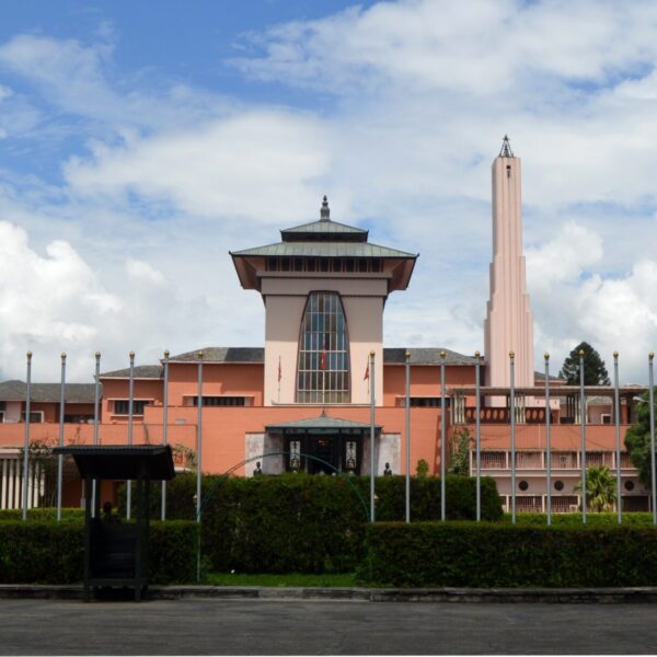 A modern architecture building with a distinctive tower and tiered roof, painted in terracotta, set against a blue sky with puffy clouds, enclosed by a fence with ornamental poles and a small bus shelter in the foreground.