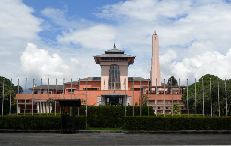A modern architecture building with a distinctive tower and tiered roof, painted in terracotta, set against a blue sky with puffy clouds, enclosed by a fence with ornamental poles and a small bus shelter in the foreground.