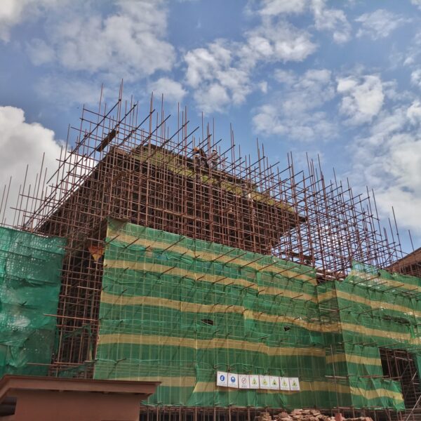 A building under construction with extensive scaffolding and green safety netting against a cloudy blue sky.