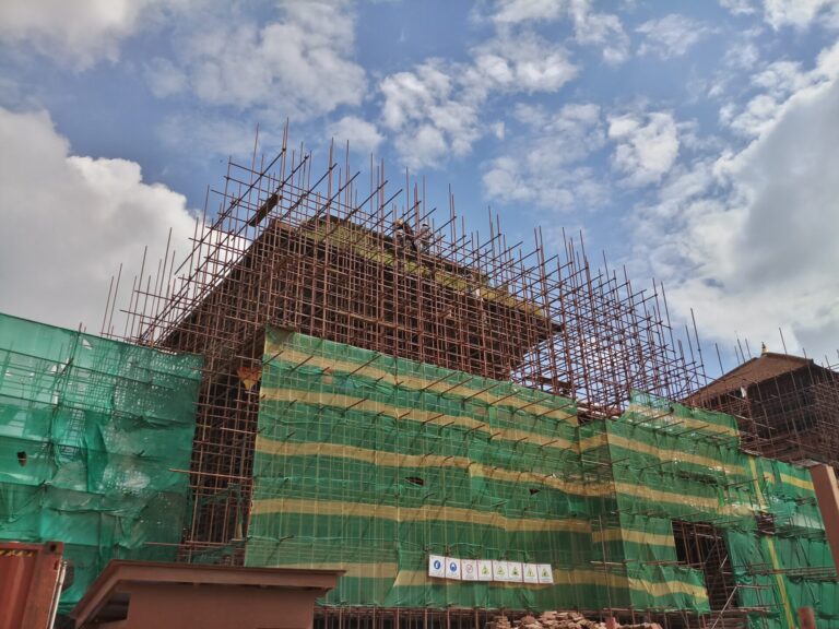 A building under construction with extensive scaffolding and green safety netting against a cloudy blue sky.