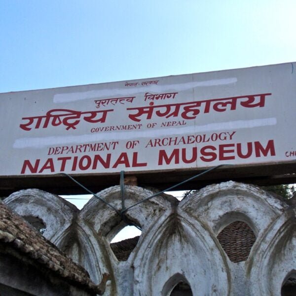 A signboard reading "Department of Archaeology National Museum, Government of Nepal" in both Nepali and English, with the name "Chhauni" at the bottom right, placed above an old, arched stone structure against a clear sky.