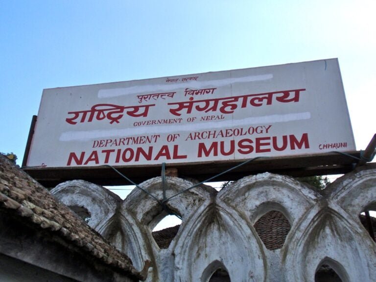 A signboard reading "Department of Archaeology National Museum, Government of Nepal" in both Nepali and English, with the name "Chhauni" at the bottom right, placed above an old, arched stone structure against a clear sky.