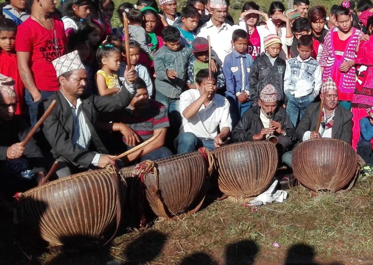 Three musicians playing traditional Nepalese drums with a crowd of onlookers in the background.