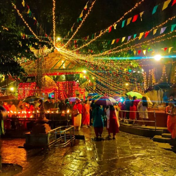 A festive outdoor scene at night with people holding candles and umbrellas under strings of lights and colorful flags.