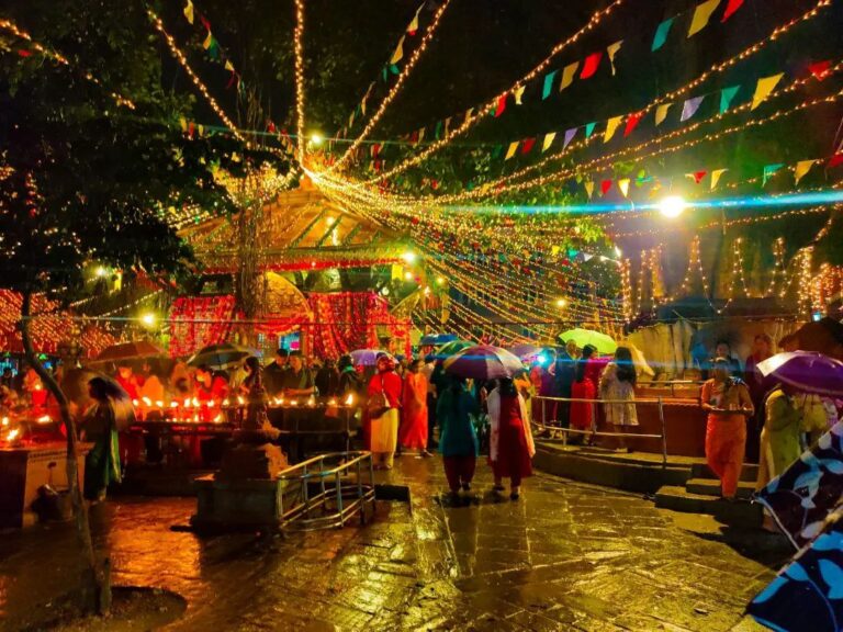 A festive outdoor scene at night with people holding candles and umbrellas under strings of lights and colorful flags.