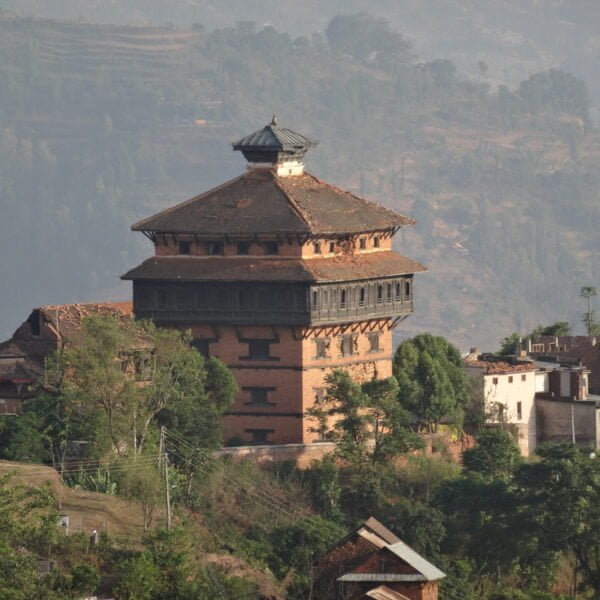 A traditional multi-storied building with a tiered pagoda-style roof stands prominently among other structures in a hilly landscape with terraced fields in the background.