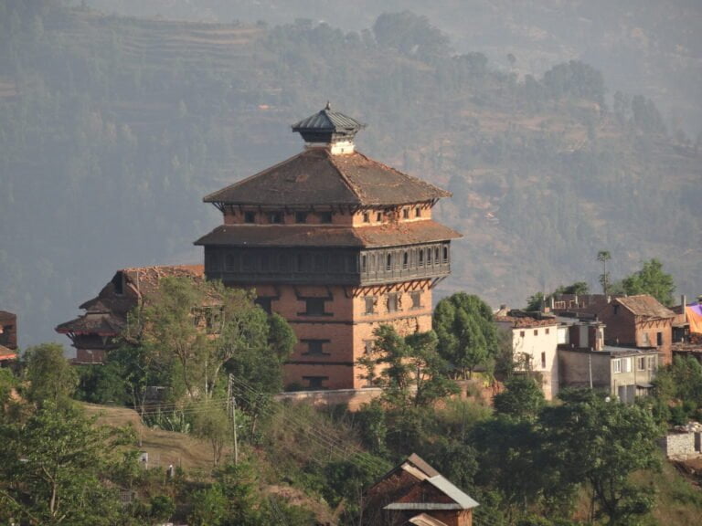 A traditional multi-storied building with a tiered pagoda-style roof stands prominently among other structures in a hilly landscape with terraced fields in the background.