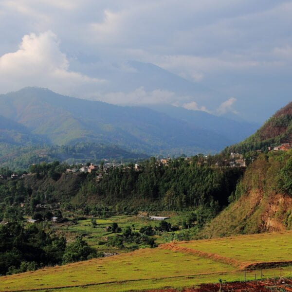 A scenic view of terraced fields in the foreground with a village nestled on the slopes of lush green hills, and misty mountains in the background under a partly cloudy sky.