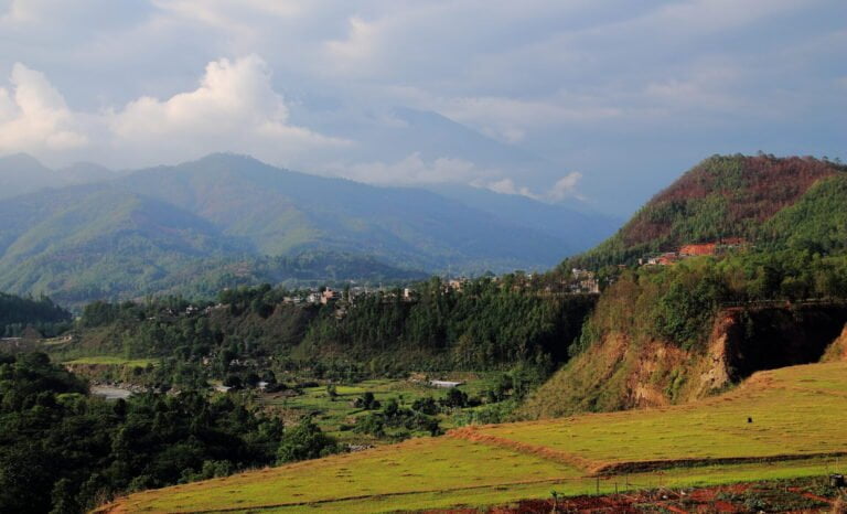 A scenic view of terraced fields in the foreground with a village nestled on the slopes of lush green hills, and misty mountains in the background under a partly cloudy sky.