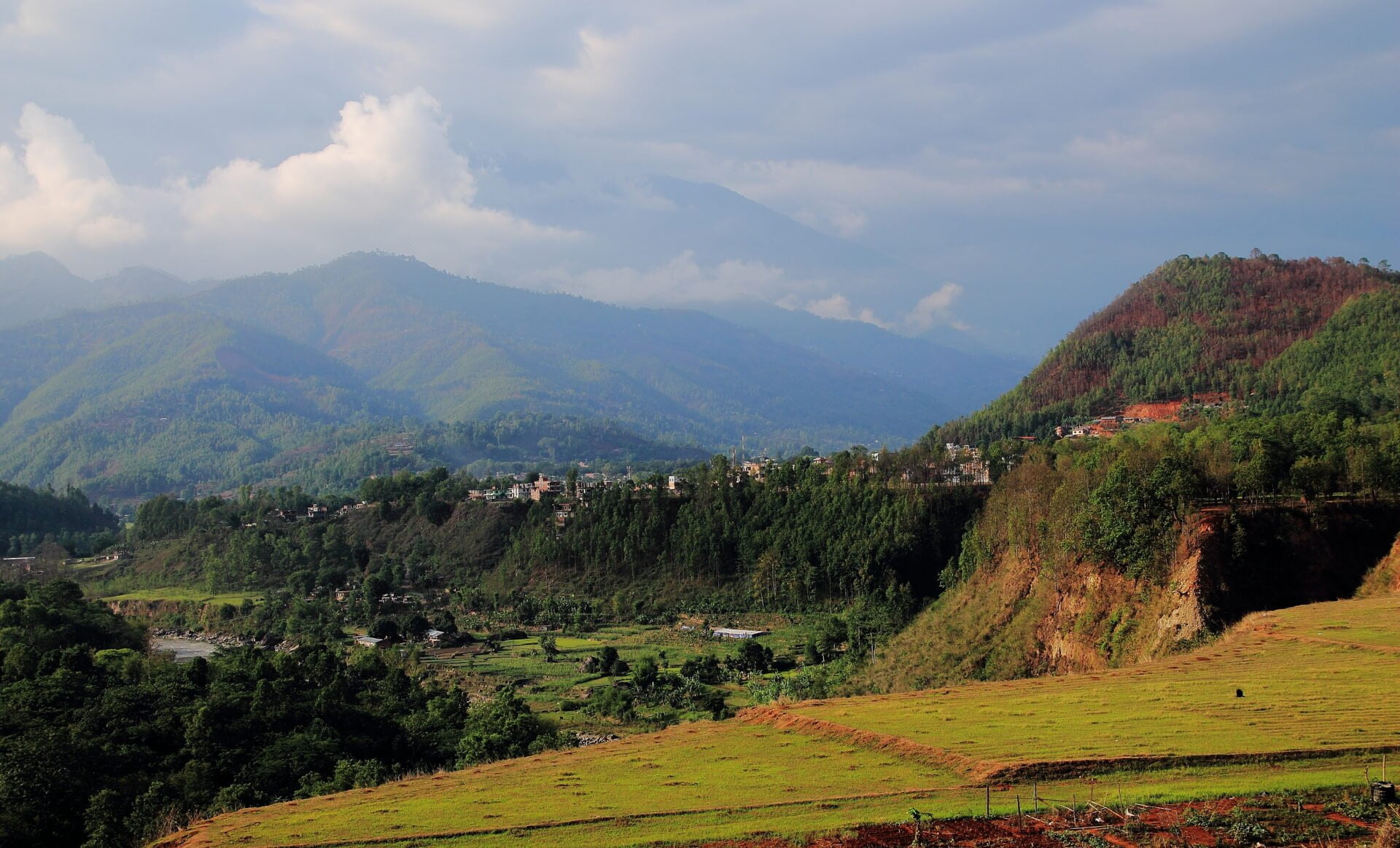 A scenic view of terraced fields in the foreground with a village nestled on the slopes of lush green hills, and misty mountains in the background under a partly cloudy sky.