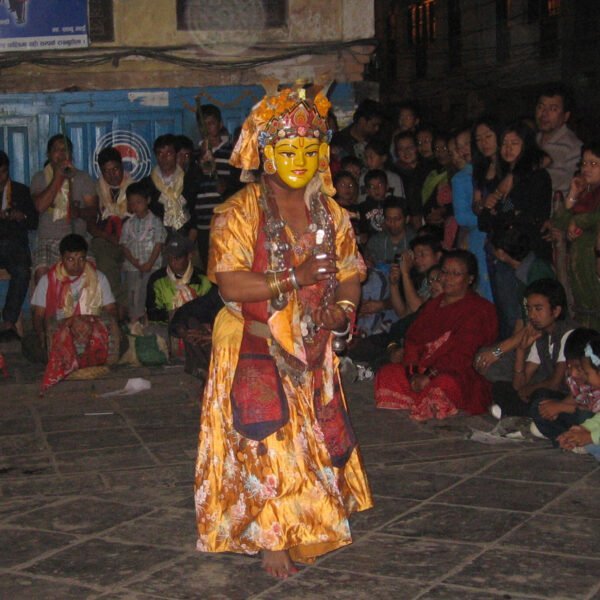 A performer in traditional costume and mask depicting a deity performs in front of an attentive audience seated on the ground at an outdoor night event.