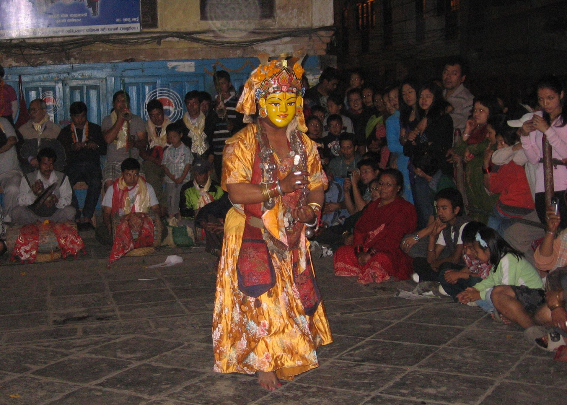 A performer in traditional costume and mask depicting a deity performs in front of an attentive audience seated on the ground at an outdoor night event.